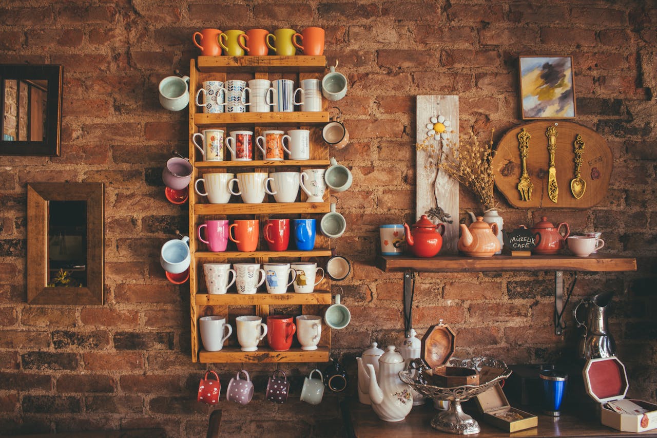 our-story A collection of colorful vintage mugs and teapots displayed on shelves against a rustic brick wall.