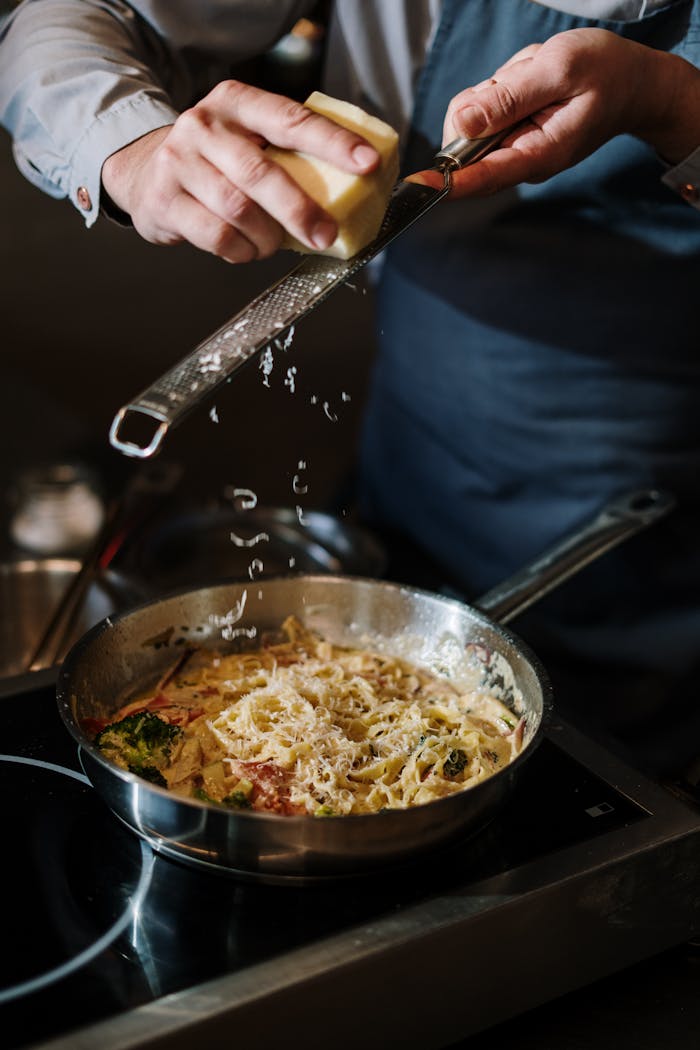 who-we-are A chef in apron grates cheese onto pasta in a modern kitchen, capturing culinary artistry in action.